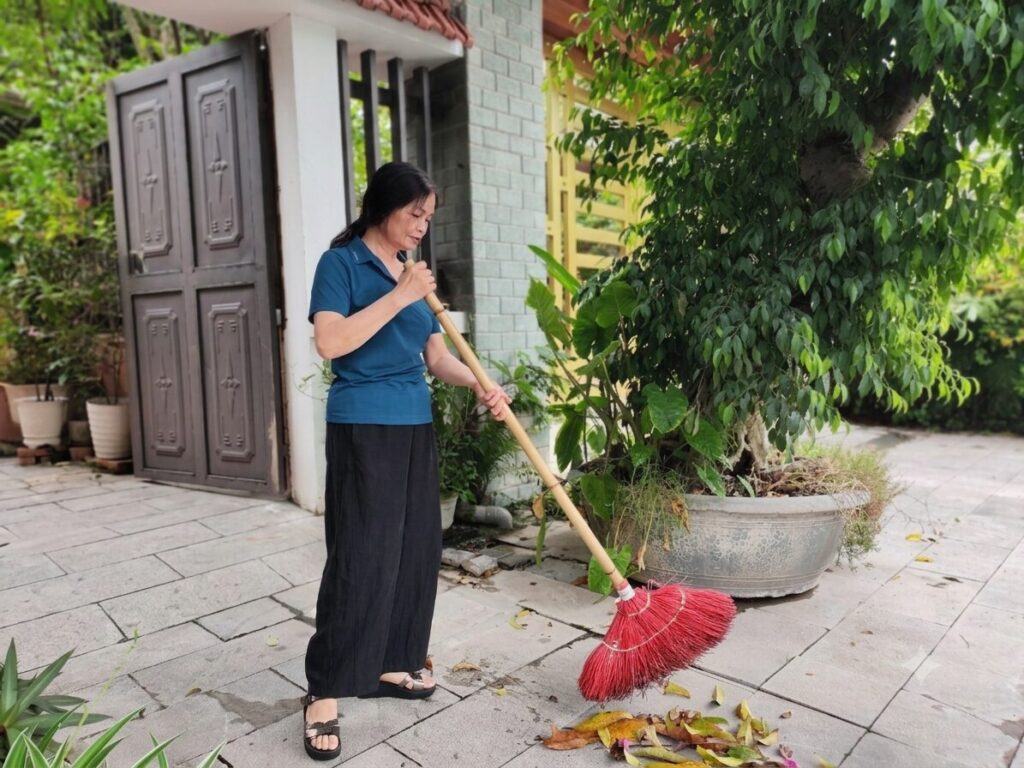 Nữ hộ sinh Nguyễn Thị Minh quét dọn sân nhà trong cuộc sống thường nhật sau khi nghỉ hưu, phản ánh nhịp sống giản dị của nhân vật trong bài viết.